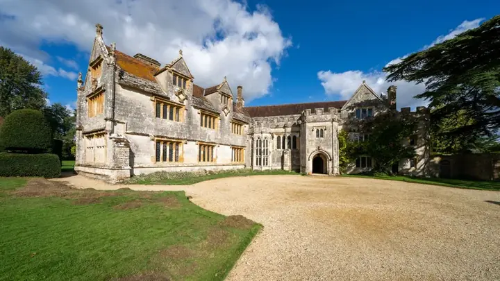 A medieval stone manor house with mullioned windows behind a gravel forecourt