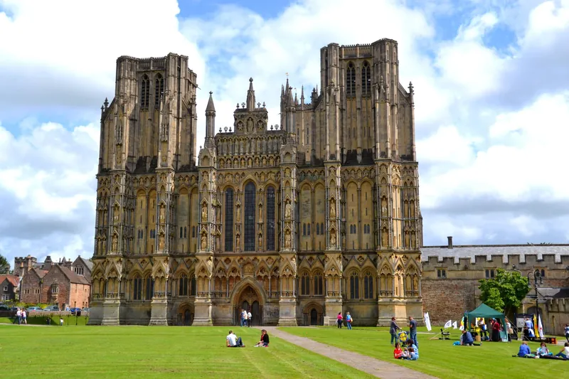 The carved west front of Wells Cathedral with visitors sitting on the green in front